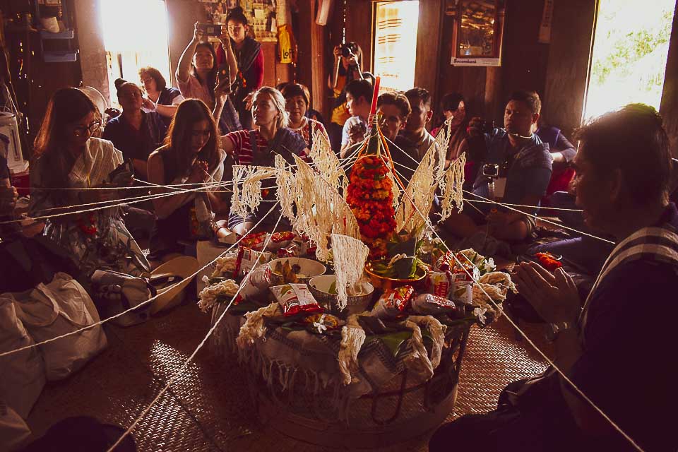 Guests gathered around a pah kwan centerpiece during a Baci ceremony, with white blessing threads draped outward to the participants
